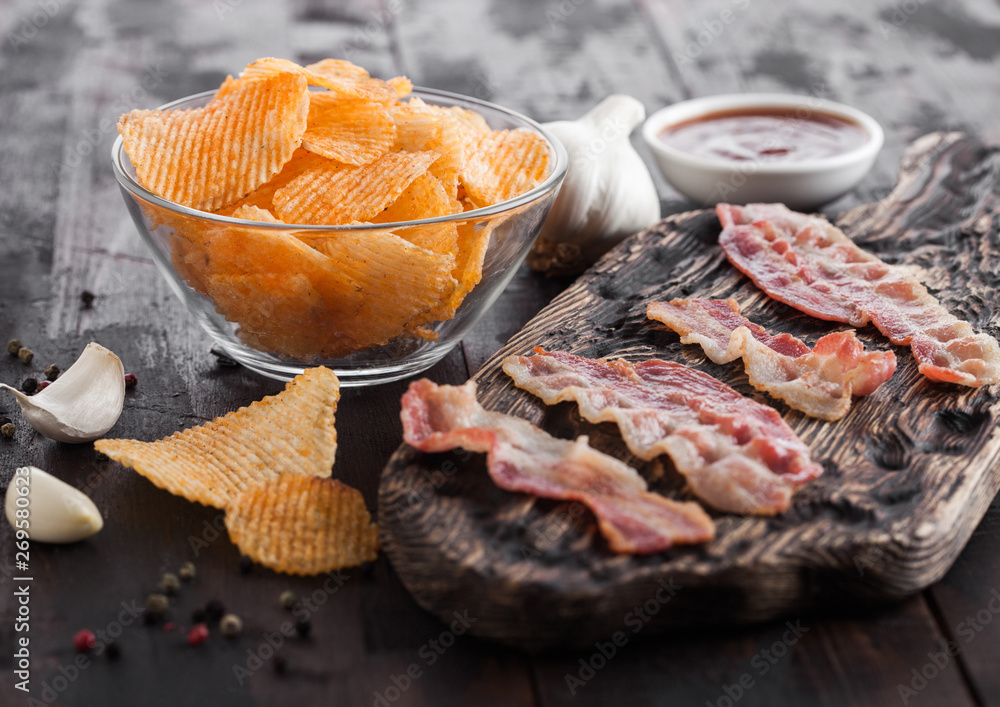Potato crisps with backon flavour with grilled bacon rashers on vintage chopping board with garlic and sauce on wooden background. Potato crisps with backon flavour with grilled bacon rashers on vintage chopping board with garlic and sauce on wooden background.