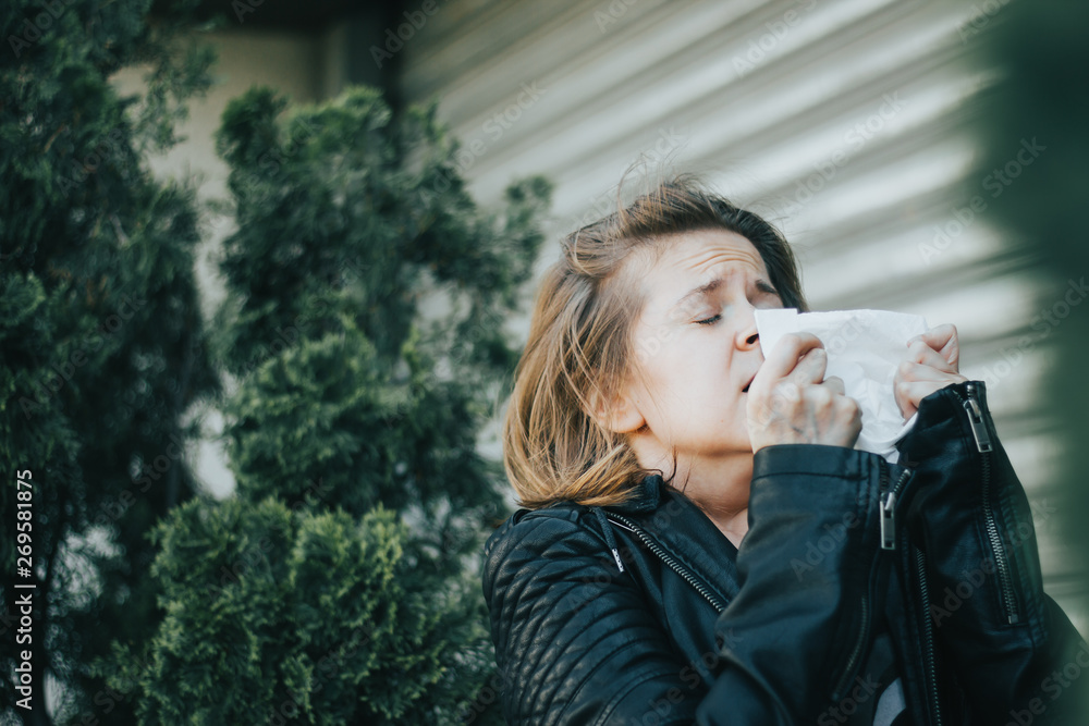 Fototapeta premium Young woman with brown hair wearing a leather jacket holding a white napkin while sneezing – Girl having an allergic reaction or a virus infection – Concept image for allergy and sickness