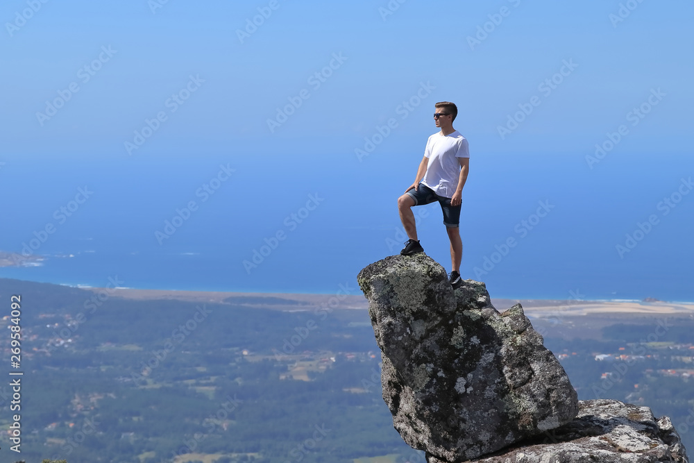 IN SUMMER TIME BOY ON VACATION ON A SUNNY DAY WITH SUNGLASSES UP ON TOP OF A ROCK WITH THE SEA AND A BEACH IN THE BACKGROUND