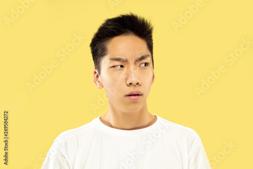 Korean young man's half-length portrait on yellow studio background. Male model in white shirt. Doubts, uncertainly, thoughtful, looking serious. Concept of human emotions, facial expression.