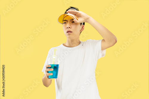 Korean young man's half-length portrait on yellow studio background. Male model in white shirt and yellow cap. Drinking cocktail. Concept of human emotions, expression, summertime, vacation, weekend. © master1305