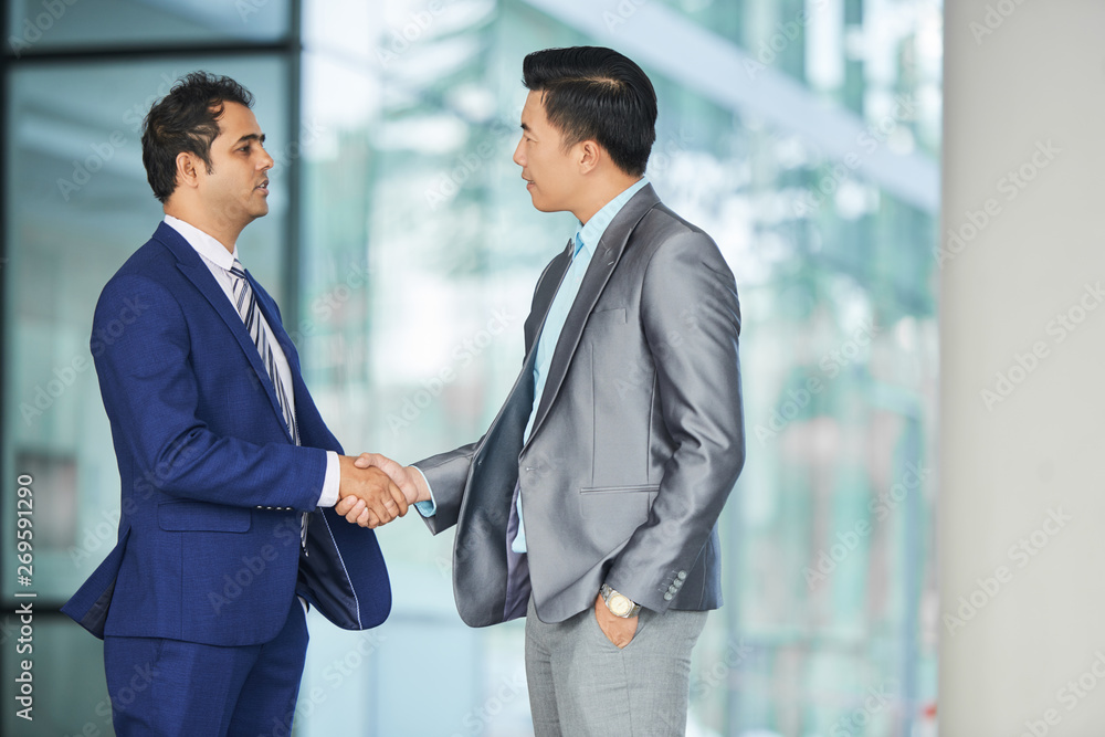Two businessmen in suits standing outdoors near the entrance in office building and shaking hands to each other Two businessmen in suits standing outdoors near the entrance in office building and shaking hands to each other