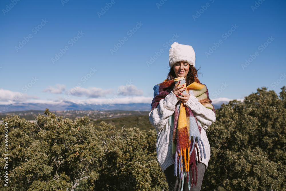 A beautiful woman is enjoying a coffee in the nature. She is stood up with an impressive view of the nature. She is wearing a colorful foulard and a white hat. She is smiling at the cup.