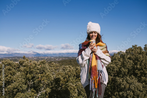 A beautiful woman is enjoying a coffee in the nature. She is stood up with an impressive view of the nature. She is wearing a colorful foulard and a white hat. She is smiling at the cup.