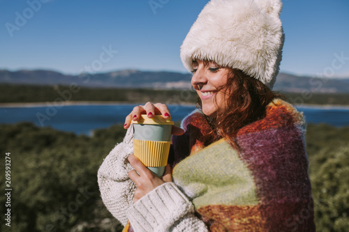 A caucasian woman is smiling while enjoying a coffee in a sunny day. She is holding the blue and yellow cup in her hands. She has dark brown hair and she is wearing a white hat and a white foulard. © NatRomero