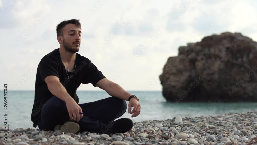 Young man with closed eyes sitting and relaxing in yoga style near amazing sea side view. Handsome guy meditating on the background of a rock and the sea. Time laps. Cyprus. Paphos Young man with closed eyes sitting and relaxing in yoga style near amazing sea side view. Handsome guy meditating on the background of a rock and the sea. Time laps. Cyprus. Paphos