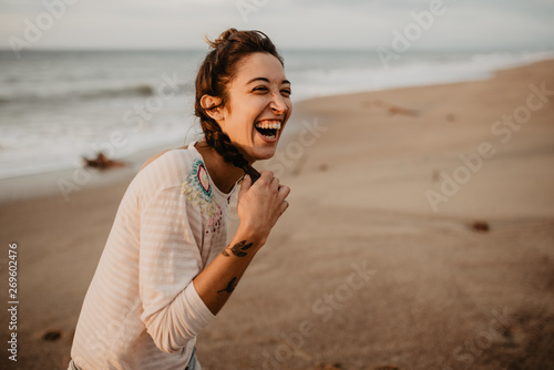 Young lady in casual outfit touching braid and laughing out loud while standing on sandy seashore