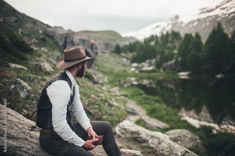 A man in a hat sits on the lake and looks at the landscape and holds a notebook in his hands A man in a hat sits on the lake and looks at the landscape and holds a notebook in his hands