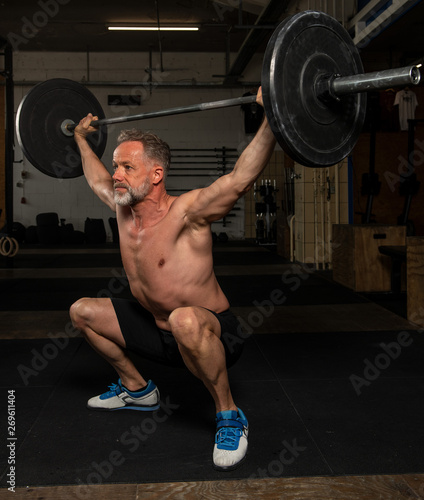 Ein durchtrainierter Mann mittleren Alters macht oberkörperfrei Overhead squats. Fitnesstraining mit der Langhantel. 