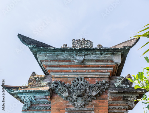 Dusun Ambengan, Bali, Indonesia - February 25, 2019: Decorated top of pillar set in wall at family compound has dark gray ornaments on red bricks under light blue sky. Some green foliage. photo