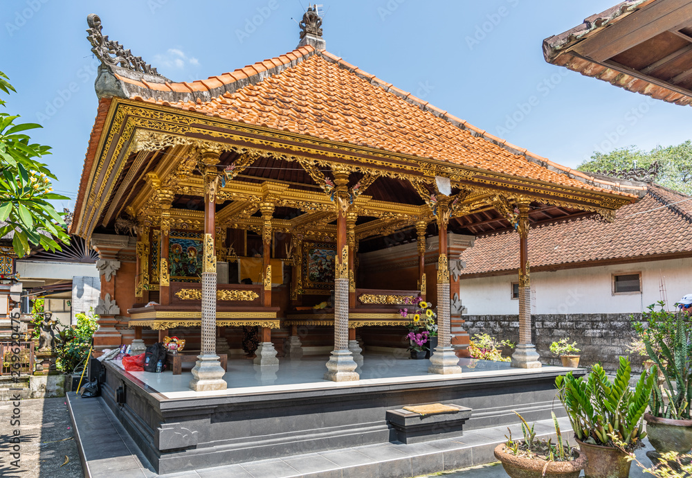 Dusun Ambengan, Bali, Indonesia - February 25, 2019: Family compound. Open wall Hundu temple with large beds in front of altar. Browns and lots of gold, blue sky, green vegetation, red roof.
