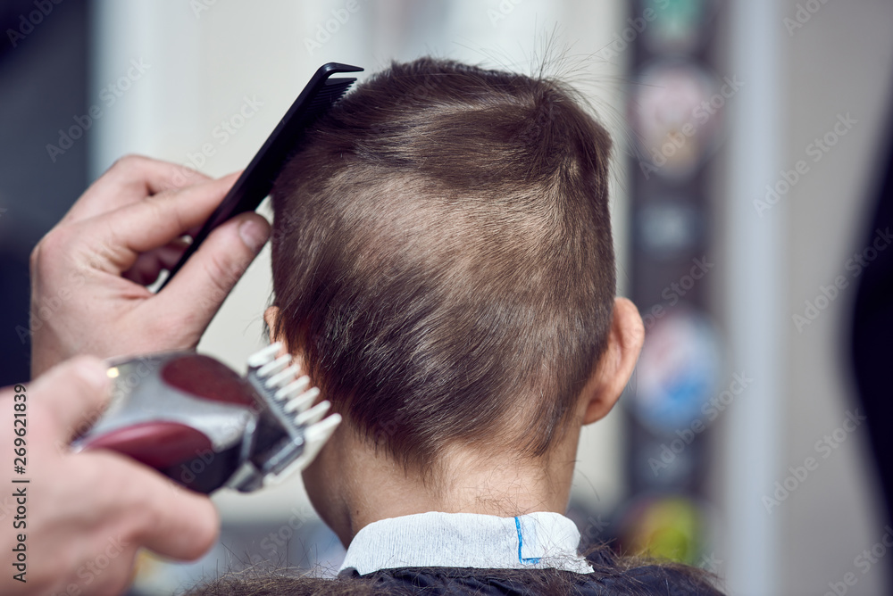 Fototapeta premium Barber making a haircut to a cute European boy using cutting machine.