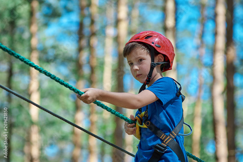 Young child boy in safety harness and helmet attached with carbine to cable moves confidently along rope way in recreation park. Sport, game, leisure concept.