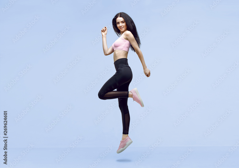 Fototapeta premium Woman in trendy sportswear jumping. Smiling beautiful slim brunette young girl in fashion leggings and pink top expressing happy emotions on blue background.