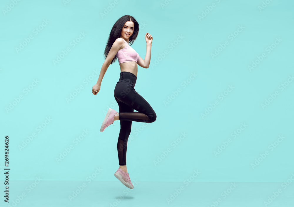Woman in trendy sportswear jumping. Smiling beautiful slim brunette young  girl in fashion leggings and pink top expressing happy emotions on blue  background. Stock Photo | Adobe Stock
