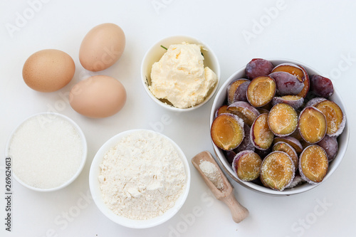 Ingredients for plum pie baking on a white background: butter, flour, sugar, baking powder, eggs, frozen plums