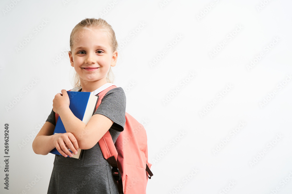Little girl photographed against white background wearing school uniform dress isolated holding a coral backpack on both shoulders and book Little girl photographed against white background wearing school uniform dress isolated holding a coral backpack on both shoulders and book