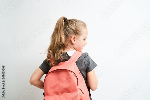 Back view of little girl photographed against white background wearing school uniform dress isolated holding a coral backpack on one shoulder