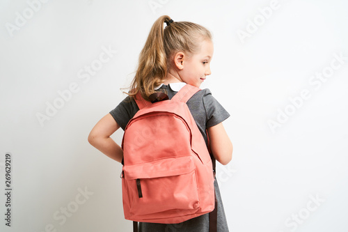 Little girl photographed against white background wearing school uniform dress isolated holding a coral backpack on both shoulders