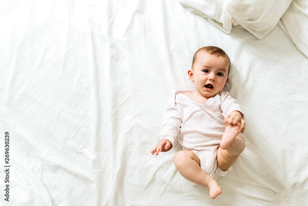 Happy and smiling adorable 6 month old baby girl lying on a bed, lifestyle isolated on natural white background. - obrazy, fototapety, plakaty 