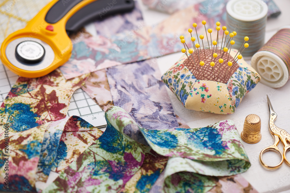Fototapeta premium Brightly colored strips of fabric, rotary cutter, pin cushion and quilting accessories on white wooden surface