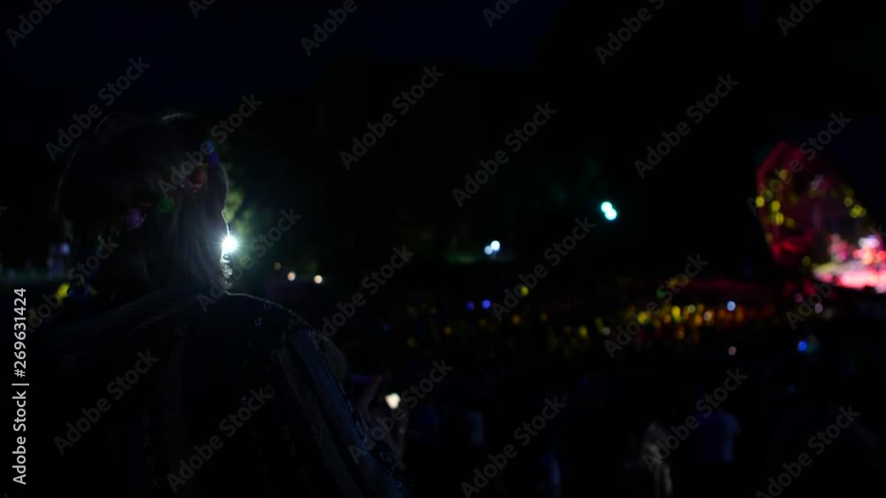 A women dancing at night with a flashing light headband on in the foreground whilst an act is playing on the main stage with a large crowd at a festival in the background. Great summer festival vibes. A women dancing at night with a flashing light headband on in the foreground whilst an act is playing on the main stage with a large crowd at a festival in the background. Great summer festival vibes.