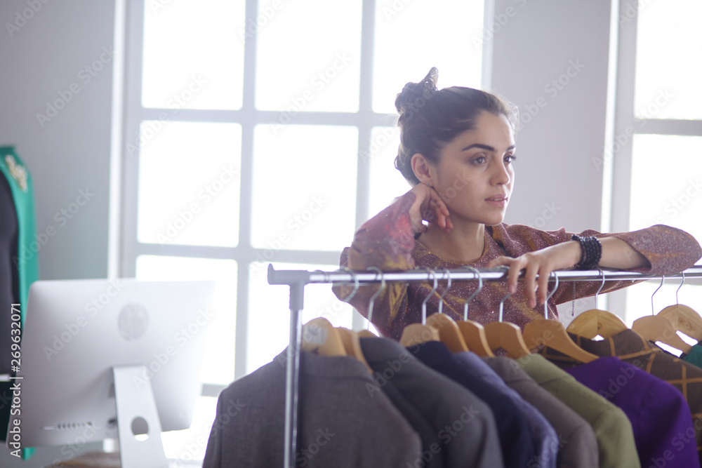 Beautiful young stylist holding clipboard near rack with designer clothes on white background Beautiful young stylist holding clipboard near rack with designer clothes on white background