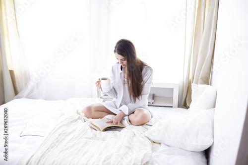 Young beautiful happy brunette woman in black silk dressing gown lying in bed, drinking morning coffee, white cup. Womens day holiday