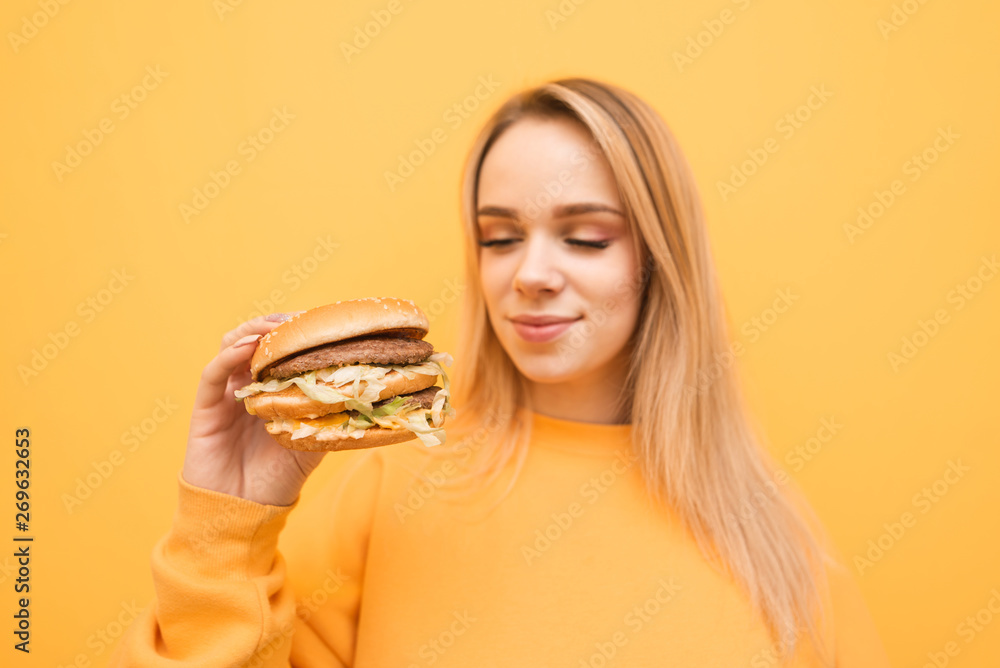 Attractive blonde with a burger in his hand stands on a yellow background, staring staring at fast food. Hungry girl holds the burger in her hands on a yellow background. Focus on the burger. Attractive blonde with a burger in his hand stands on a yellow background, staring staring at fast food. Hungry girl holds the burger in her hands on a yellow background. Focus on the burger.