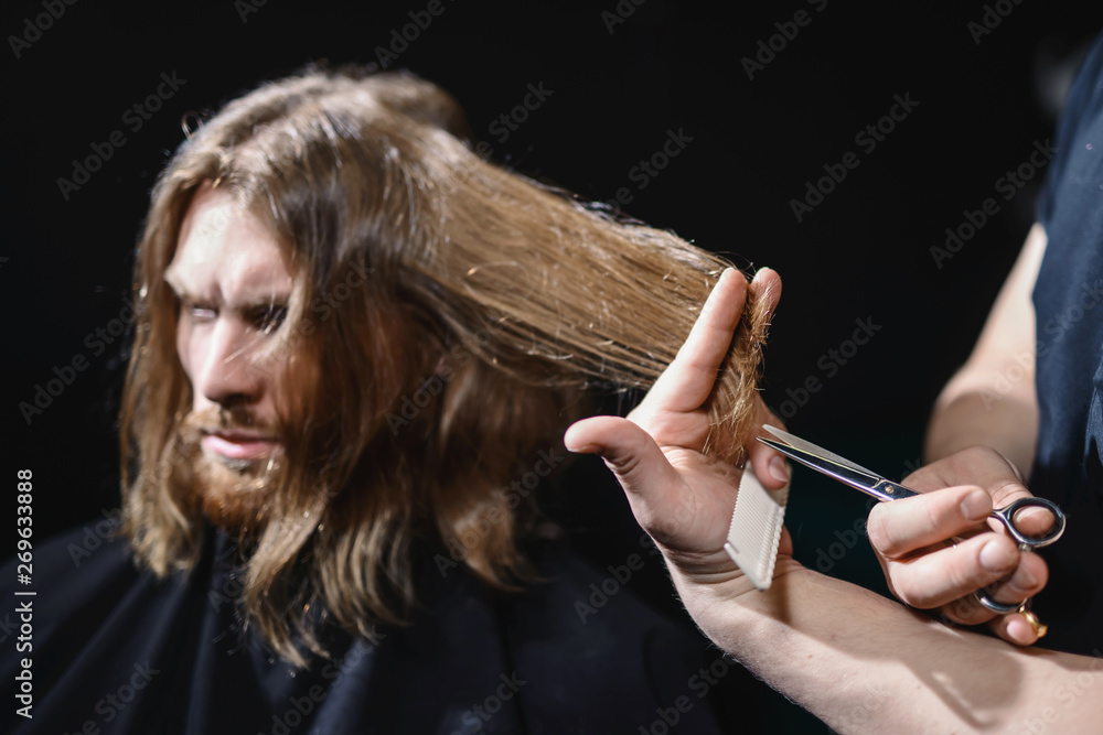 Obraz premium Handsome bearded man, hipster with beard and moustache has haircut in hairdressing saloon or barbershop. Barber works with hair trimmer or clipper. Black background isolated