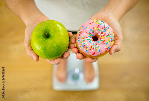 Woman on scale measuring weight holding apple and donuts choosing between healthy or unhealthy food photo