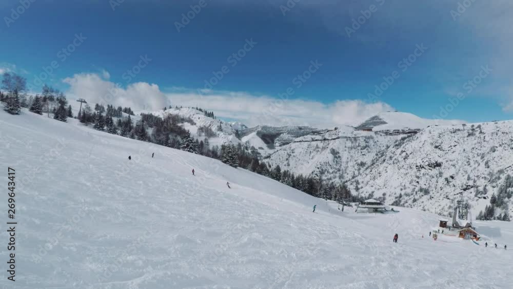 Mountain ski resort in winter with skiers enjoying a sunny day on the slopes. People skiing downhill toward a ski lodge and the lift station. Aerial view from drone moving uphill. Mountain ski resort in winter with skiers enjoying a sunny day on the slopes. People skiing downhill toward a ski lodge and the lift station. Aerial view from drone moving uphill.