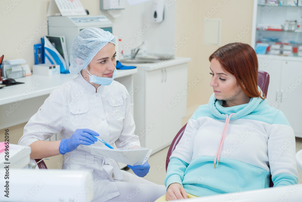 Young beautiful female dentist talking to a patient in the office. A red-haired woman sits in a chair with an orthodontist and listens to appointments. Dental services Young beautiful female dentist talking to a patient in the office. A red-haired woman sits in a chair with an orthodontist and listens to appointments. Dental services
