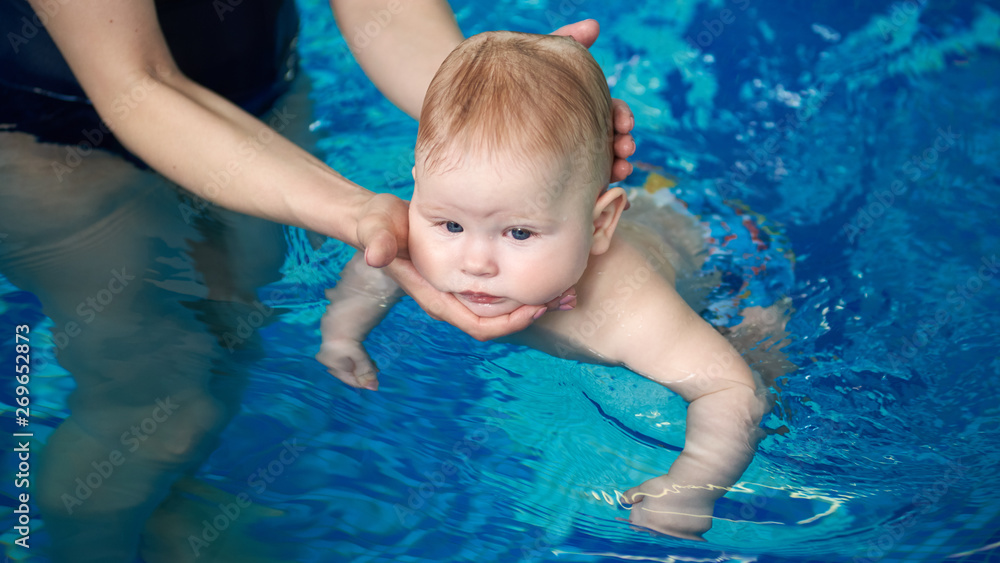 Active baby swimming in woman's hands in swimming pool. Cropped female hands holding carefully baby's head. First attempt to independently swimming for harmonious development. Blue water on background