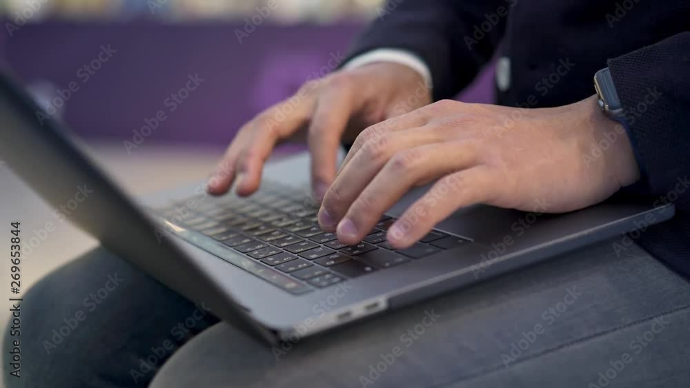 Handheld close up shot of male hands with smartwatch typing on grey keyboard of modern laptop lying on knees. Close up of young businessman hands dressed in suit typing on keyboard Handheld close up shot of male hands with smartwatch typing on grey keyboard of modern laptop lying on knees. Close up of young businessman hands dressed in suit typing on keyboard