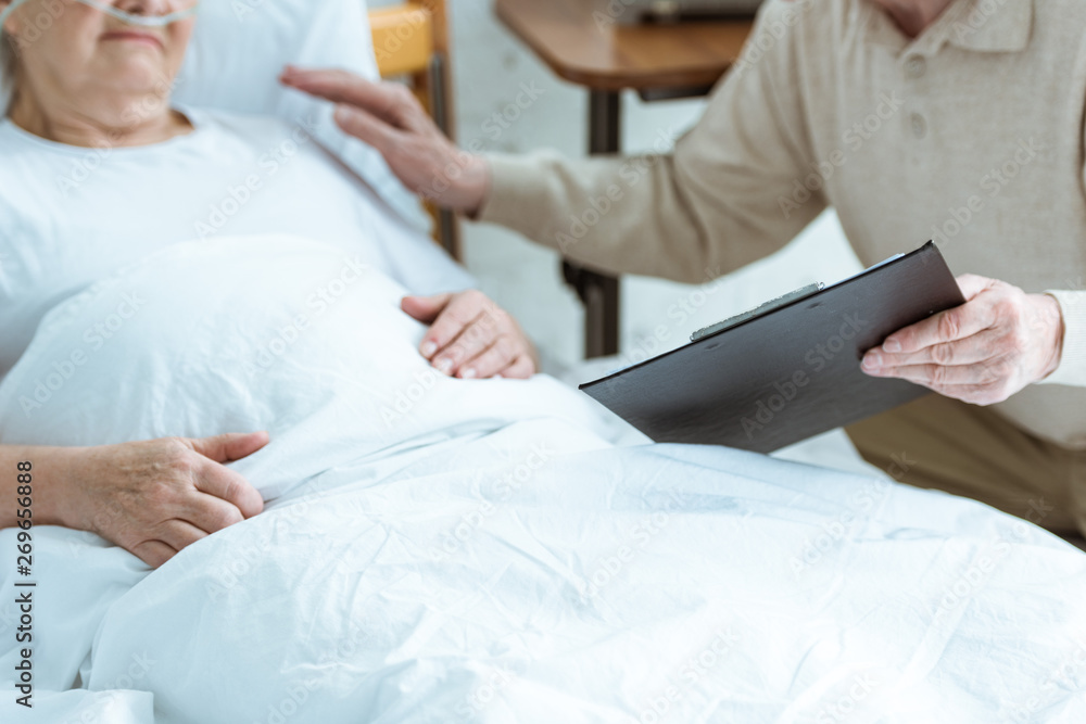 Obraz premium cropped view of senior man holding clipboard with sick wife in clinic