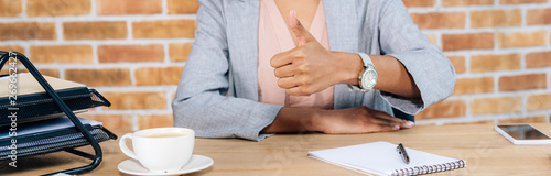 panoramic shot of Casual african american businesswoman at desk with coffee and notebook showing thumb up