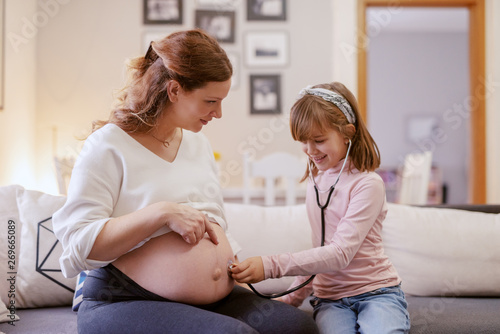 Beautiful Caucasian pregnant mother sitting in living room with her daughter. Daughter listening with stethoscope baby's heartbeats. Living room interior.