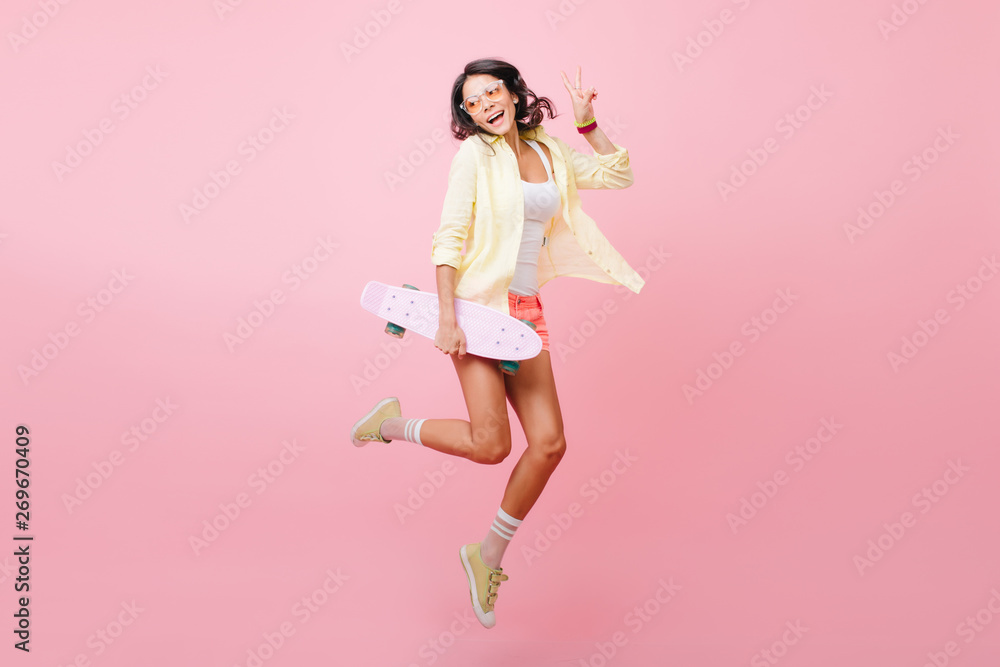 Full-length portrait of joyful dark-haired girl jumping with skateboard on pink background. Amazing hispanic lady in colorful attire dancing in studio holding longboard and laughing. Full-length portrait of joyful dark-haired girl jumping with skateboard on pink background. Amazing hispanic lady in colorful attire dancing in studio holding longboard and laughing.