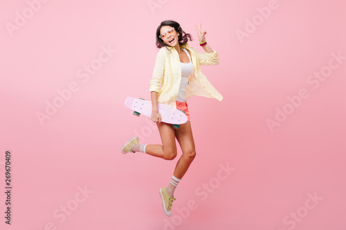 Full-length portrait of joyful dark-haired girl jumping with skateboard on pink background. Amazing hispanic lady in colorful attire dancing in studio holding longboard and laughing.