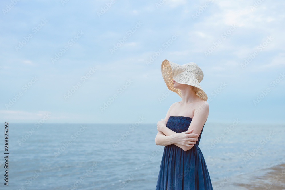 Caucasian beautiful stylish blonde woman in blue dress and in straw hat standing near the sea Caucasian beautiful stylish blonde woman in blue dress and in straw hat standing near the sea