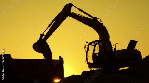 Silhouette of an excavator that loads sand into a truck at sunset. Concept construction and heavy industry, machine will be used in heavy industry business. Slow motion footage.