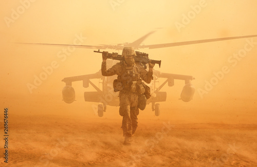 Military soldier walking at desert with gun on his shoulder in front of helicopter in sand storm. photo