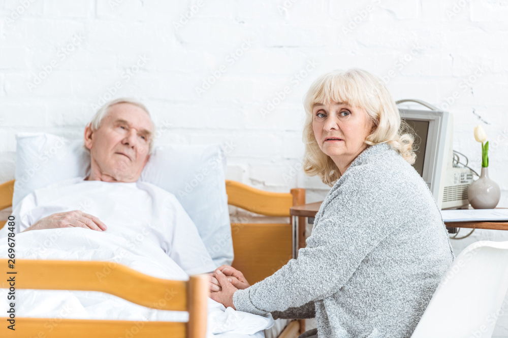 Fototapeta premium worried senior woman sitting near ill husbend and holding his hand in hospital