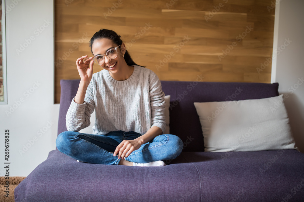 Obraz premium Beautiful young cheerful woman in glasses looking at camera with smile while sitting on the couch at home