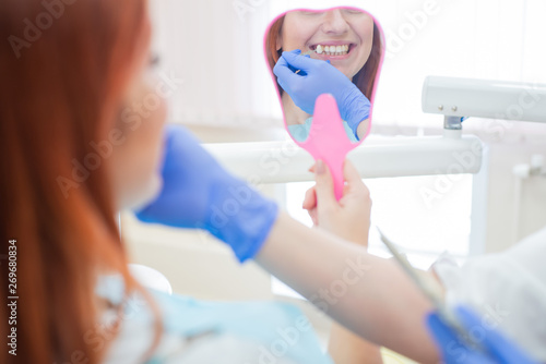 Check and select the color of the teeth in the dentist chair. Dentist makes the process of treating a beautiful young red-haired girl. Selection of a tooth implant.The reflection in the mirror.