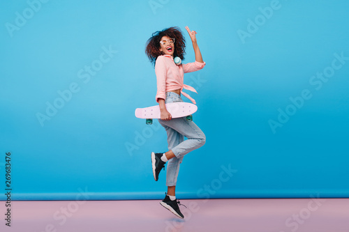 Fototapeta Naklejka Na Ścianę i Meble -  Indoor portrait of joyful girl in pink shirt standing on one leg and laughing on blue background. Fashionable curly female model with skateboard dancing during photoshoot.