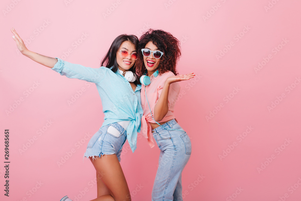 Cute hispanic girl in blue shirt embracing african female friend in studio. Lovable brunette ladies hugging on pink background with laugh. Cute hispanic girl in blue shirt embracing african female friend in studio. Lovable brunette ladies hugging on pink background with laugh.