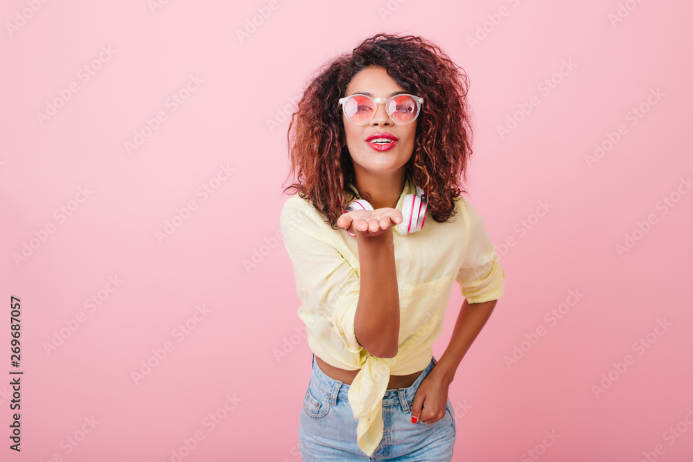 Sensual sporty woman in street outfit sends air kiss on pink background. Indoor photo of attractive female model in yellow shirt and cool sunglasses standing with lovely face expression in studio. Sensual sporty woman in street outfit sends air kiss on pink background. Indoor photo of attractive female model in yellow shirt and cool sunglasses standing with lovely face expression in studio.