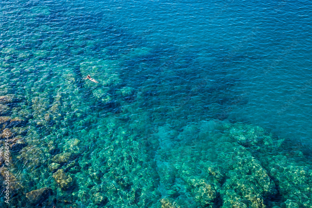 From the Cinque terre trail aerial view of the transparent, emerald, blue sea and the young woman floating in it. From the Cinque terre trail aerial view of the transparent, emerald, blue sea and the young woman floating in it.